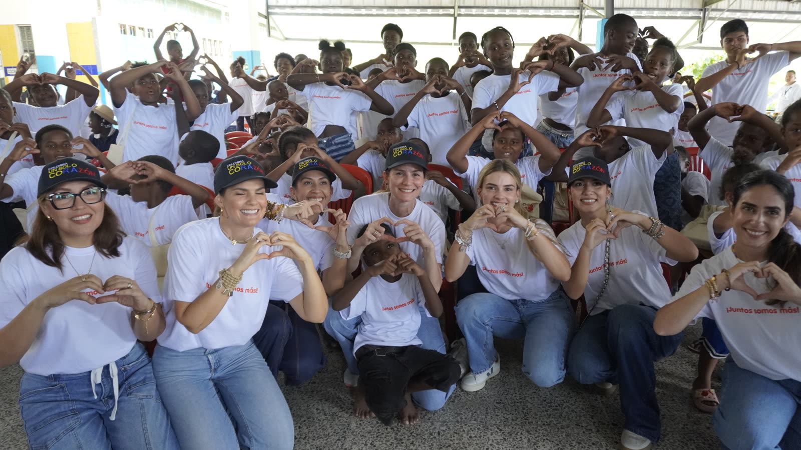 Niñas, niños y el equipo de la jornada forman corazones con las manos, con camisetas de «¡Juntos somos más!» y gorras de «Colombia, un solo corazón»